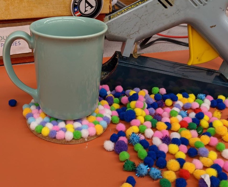 Pictured: A coaster made of small pom poms and a cork base.