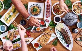 Several hands holding chopsticks are seen reaching for a variety of Japanese food items plated on white dishes.