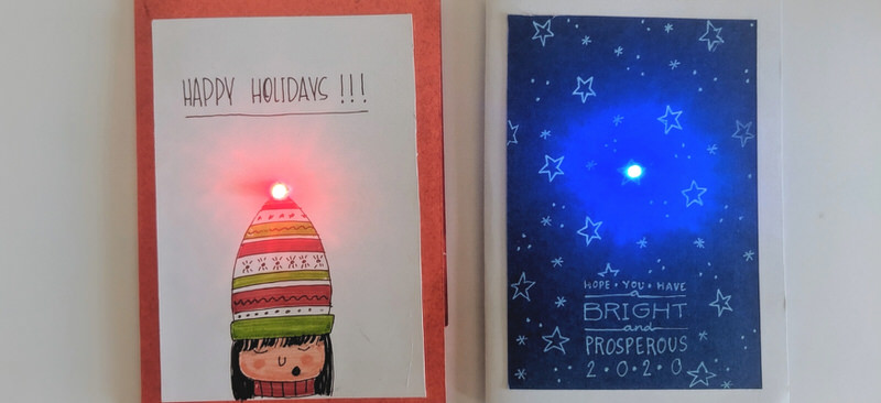 Two homemade holiday cards are side by side. One has a drawing of a person in a stiped winter hat witha glowing red light at the tip. It says, "HAPPY HOLIDAYS!!!" The other has a drawing of five-point stars in a blue sky, one of which has a glowing light. It says, "Hope you have a BRIGHT and PROSPEROUS 2020."