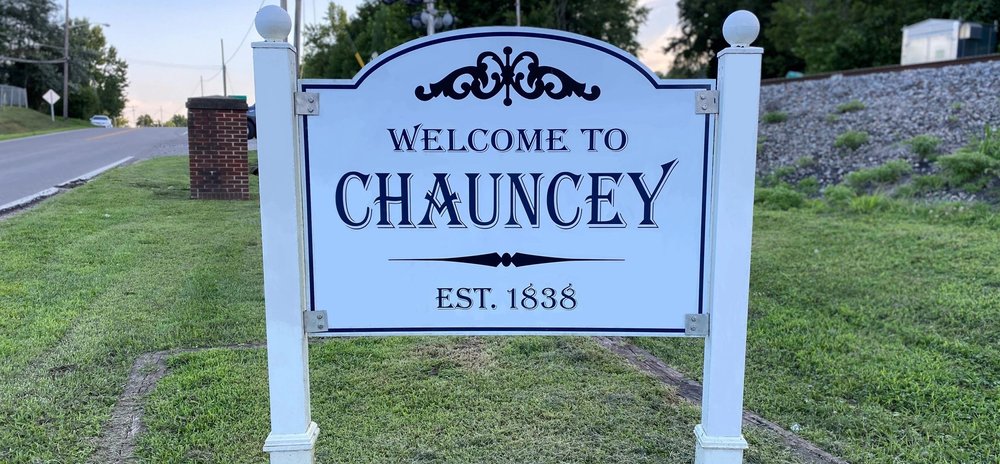 A white sign reads, "Welcome to Chauncey, est. 1838." In the background are grass, a road, and a hill covered in gravel. 