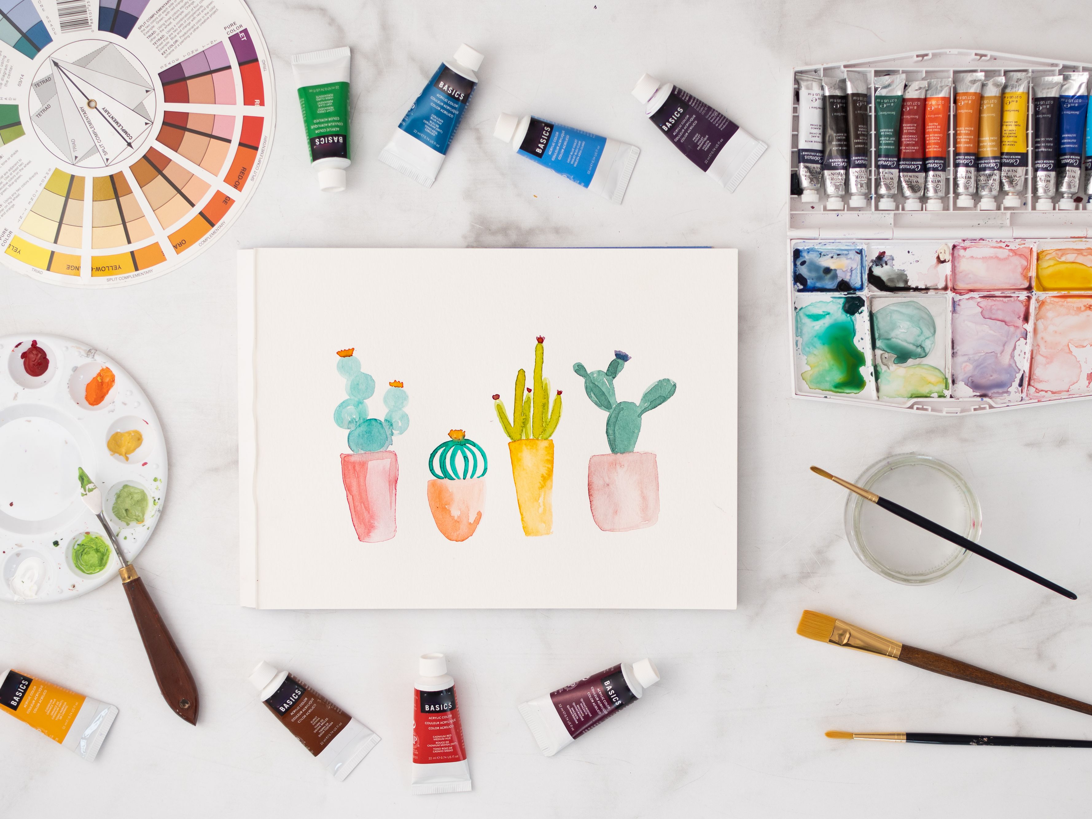 A canvas painting of four cacti is surrounded by a color wheel, a palette, paintbrushes, and tubes of paint on a white background.