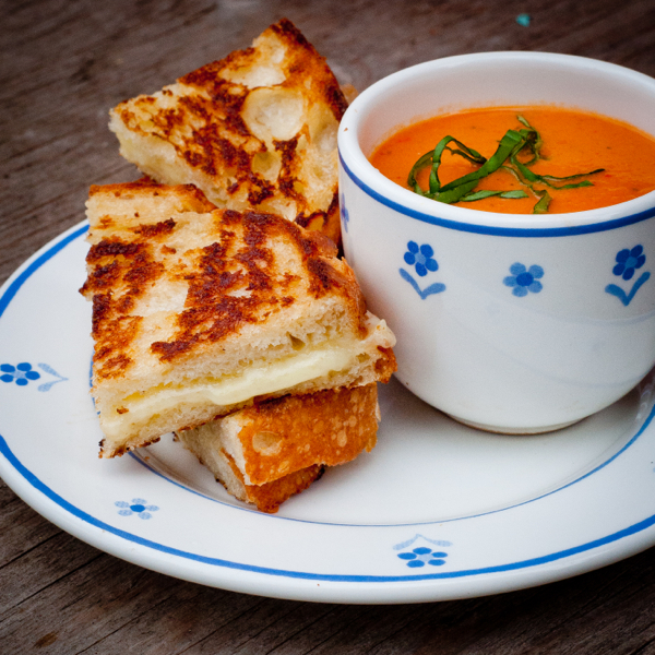A cup of tomato soup garnished with strips of basil sits beside grilled cheese cut into triangles on a plate. The plate and the cup are white with small blue flowers.