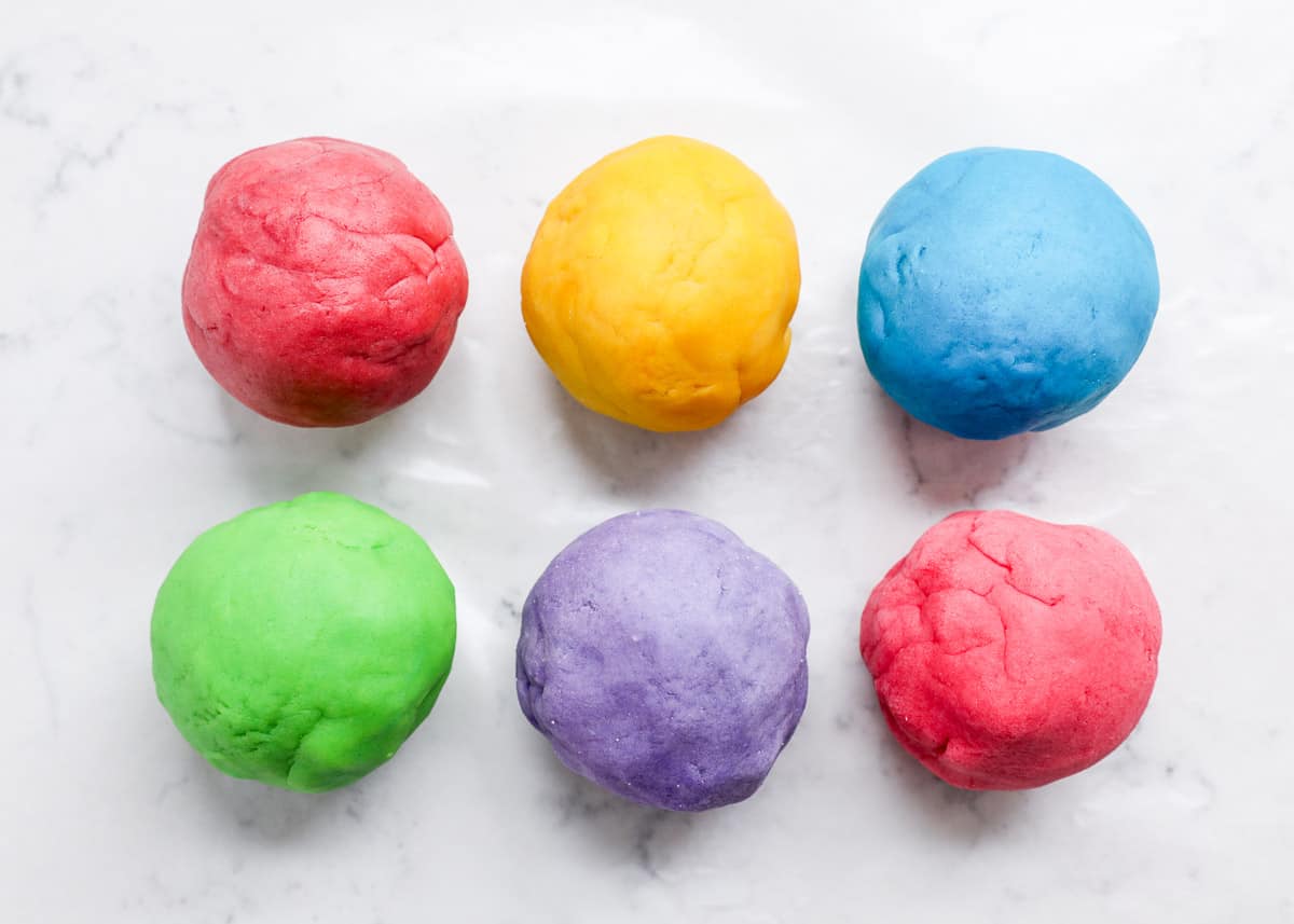 Six playdough spheres in red, yellow, blue, green, purple, and pink rest on a white marble slab.