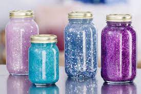 Four mason jars filled with glittery liquid-- one pink, one light blue, one dark blue, and one purple-- sit atop a smooth silver surface.