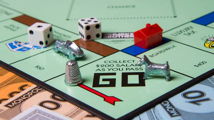 The "Go" corner of a Monopoly board hosts a dog and a thimble. Elsewhere on the board, one can see a hotel, a car, and two dice. Monopoly money lines the periphery of the board.