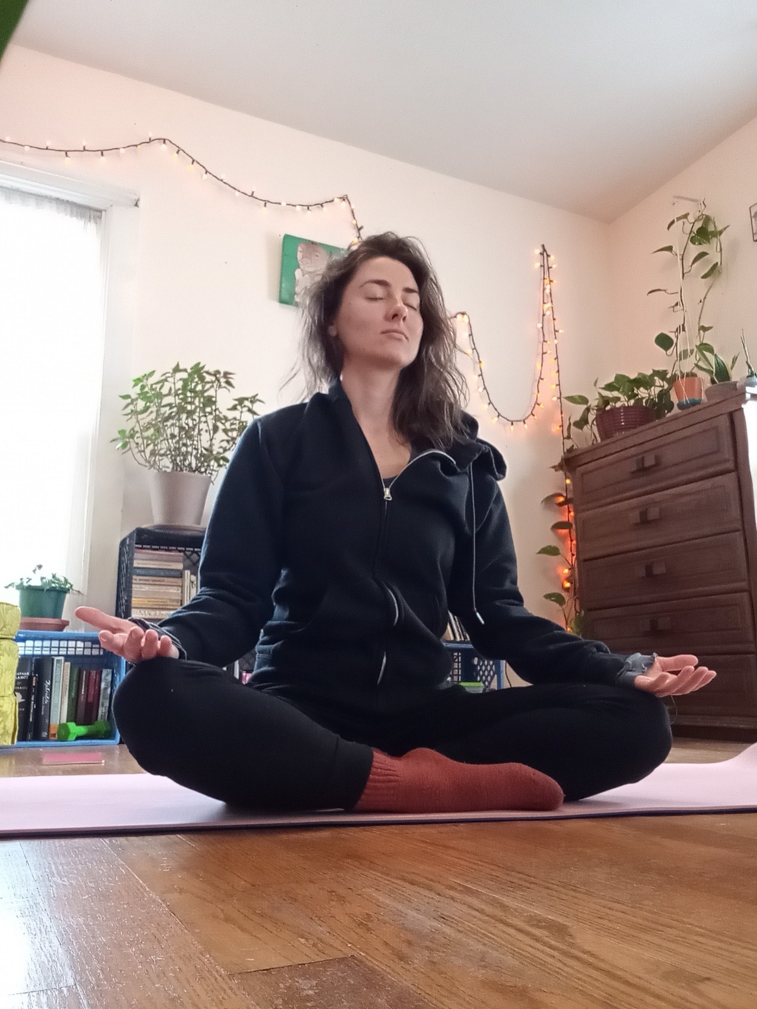 Dressed in a black zip-up hoodie, black pants, and red socks, yoga instructor Amber sits cross-legged on a red yoga mat with her hands palm-up on her knees and her eyes closed. Plants, books, and a string of white lights can be seen behind her.
