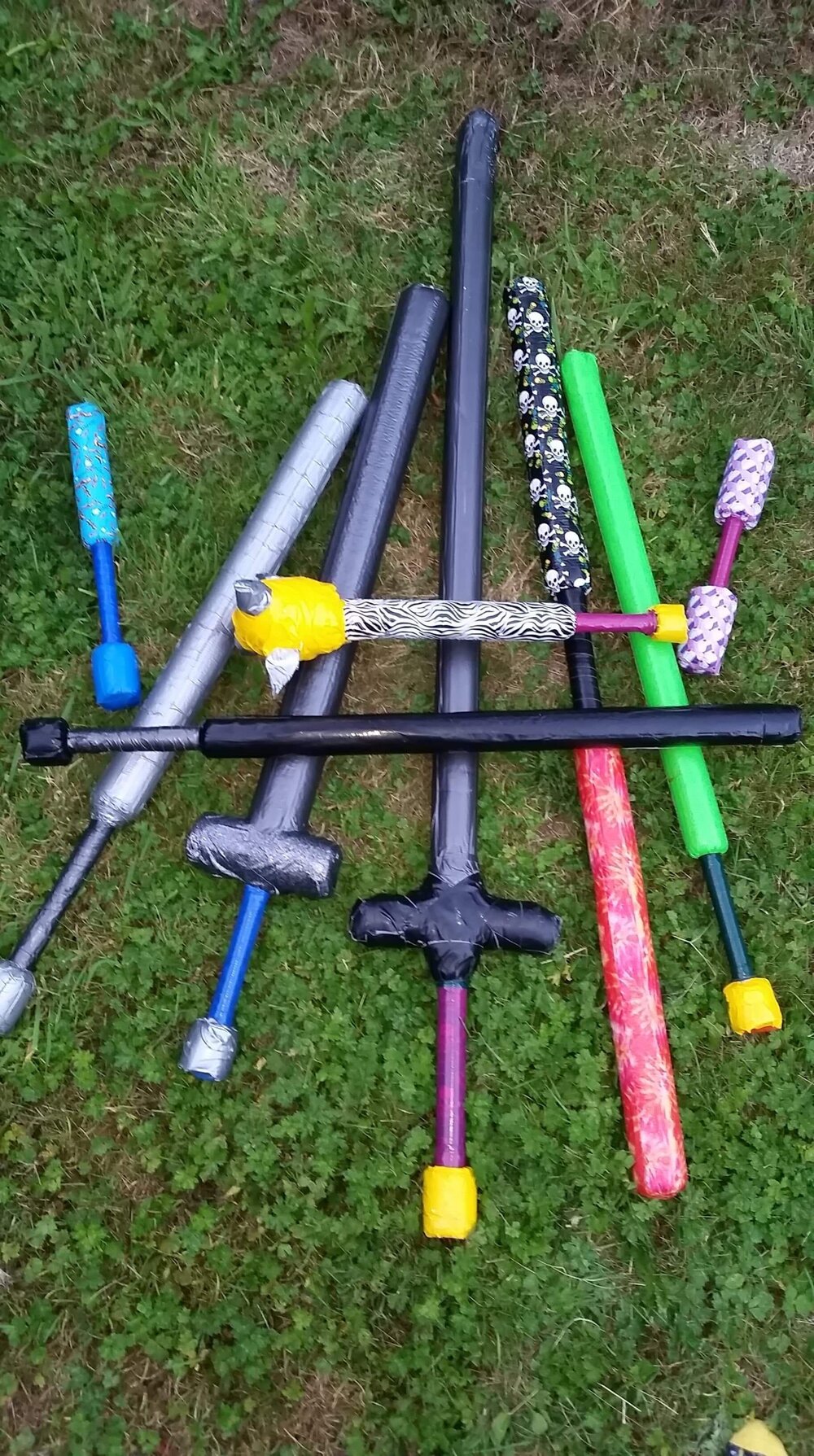 Nine duct-taped boffer swords of various lengths and colors lie in a pile on a grass lawn. 