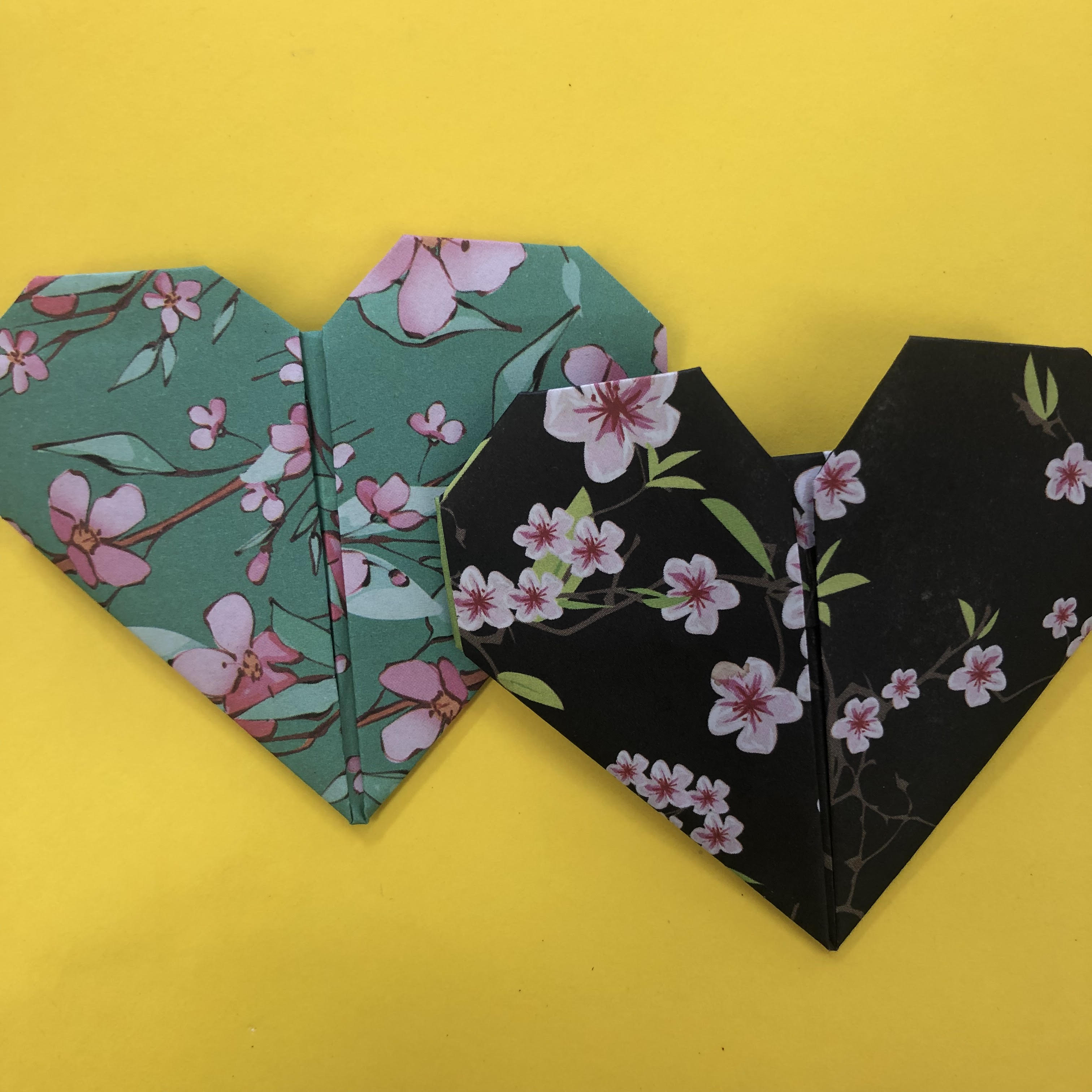 Two origami hearts on a yellow background. One is jade green with pink flowers, and one is black with pink flowers.