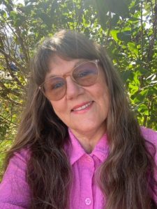 Instructor Cheryl Cesta is smiling at the camera, wearing a pink blouse and tinted glasses. She has long brown hair. There is a deciduous tree in the background.