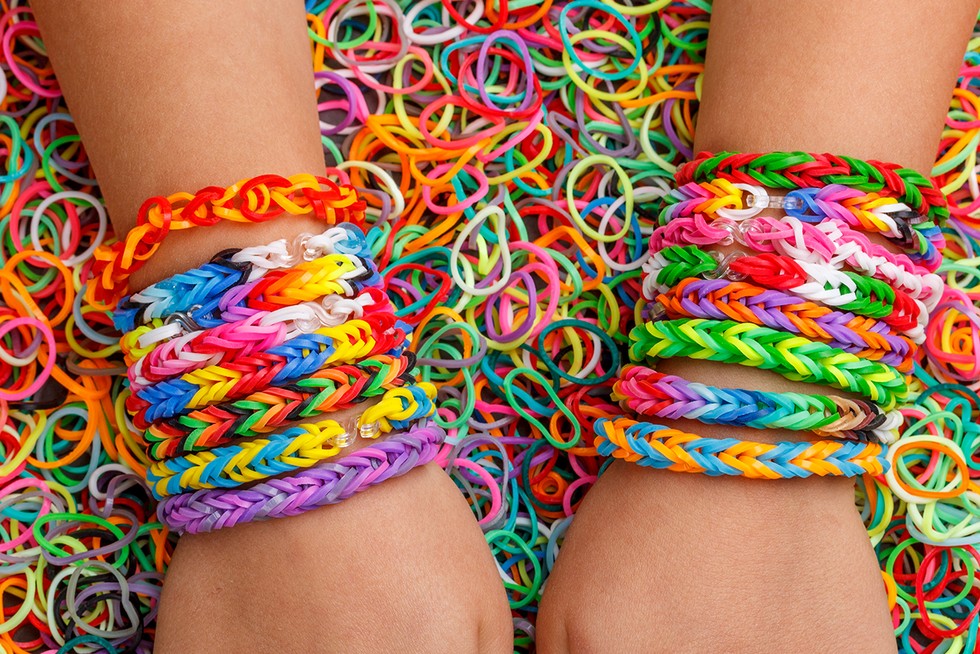 Two wrists don eight multicolored loom bracelets each. The wrists are laying on a pile of the small colorful rubber bands used to make the bracelets.