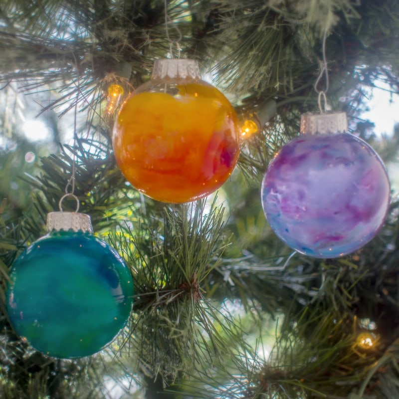 A close-up of a coniferous tree with three melted crayon ornaments hanging from it: blue, orange, and purple.