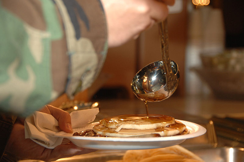 A person's hand protruding from their camoflage-patterned sleeve uses a silver ladle to scoop syrup onto a stack of pancakes.