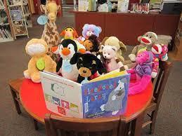 A plush cat, giraffe, penguin, dog, and several bears and monkeys are gathered around an open picture book on top of a red table.