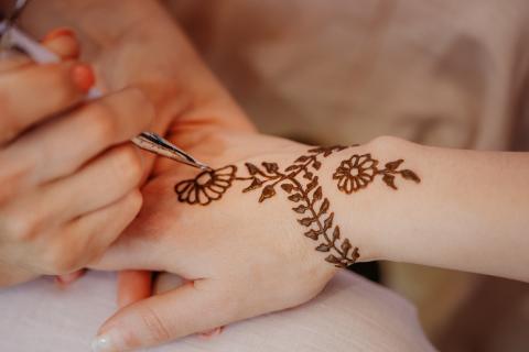 A close-up of one person drawing a flower and leaf henna tattoo on another person's hand