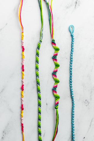 Four thin friendship bracelets made of multicolored thread lie parallel on a white marble surface.