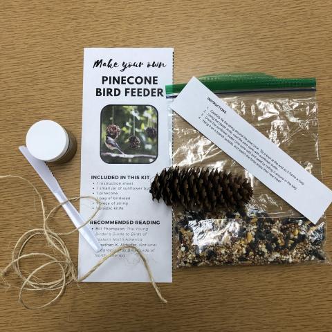 A small jar of sunflower butter, a piece of twine, a pinecone, a bag of birdseed, a plastic knife, and an inventory sheet on a wooden table