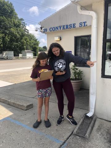 A kid and a teen stand in front of the library with clipboards, conspiring about their next move in the scavenger hunt.