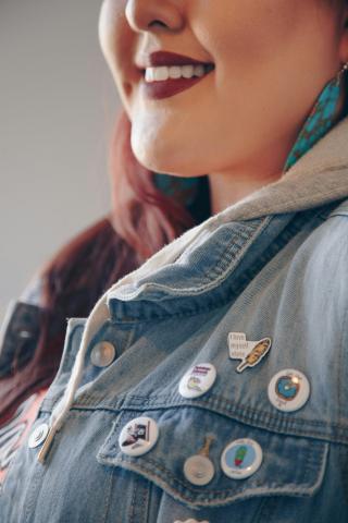Profile view of the bottom half of a smiling person's face and their chest. They are wearing a denim jacket with several buttons, one of which has an ear of corn and says "I love myself elote." The person has light brown skin, long auburn hair, dangly turquoise earrings, and dark red lipstick.