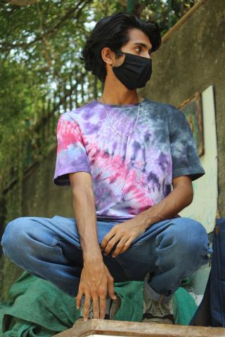 A person with brown skin and medium-length black hair squats in front of a concrete wall. They are wearing jeans; a pink, blue, and purple tie dye t-shirt; a black facemask; and a gold chain necklace.