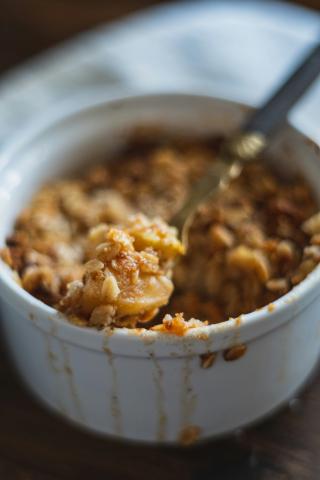 A white ramekin filled with baked apples covered in oat crumble, with a spoon protruding.