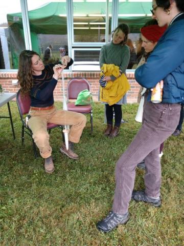 A picture of a presenter, Sasha Sigetic, using a drop spindle to spin wool while workshop participants observe.