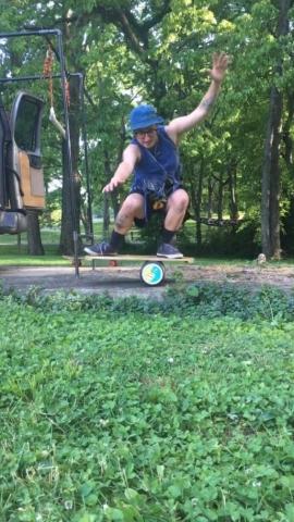 Event presenter Zest balances on a plank on top of a wheel. He is wearing a bucket hat, a tank top, and shorts, all navy blue. Grass is in the foreground and trees are in the background.