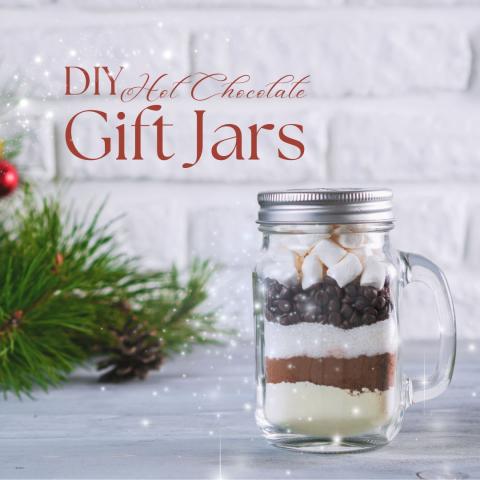 A jar full of instant hot chocolate ingredients sits in front of a white brick wall. Pine needles lay beside it. In the top left corner, script reads "DIY Hot Chocolate Gift Jars."