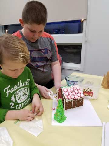 Two children make a gingerbread house on a yellow table. Tubs of candy and ice cream cones are in the background.