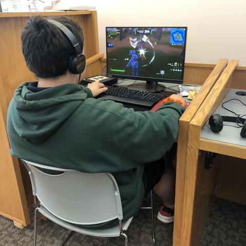 A teenager sits at a library computer and plays Fortnite. They have short, dark brown hair and are wearing a green hoodie and headphones.