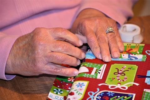 A person in a pink sweater wraps a rectangular prism in Christmas wrapping paper. A roll of tape is nearby.