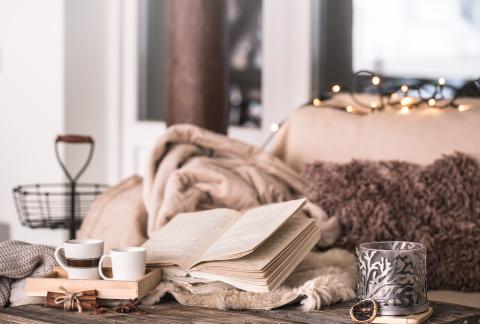 Still life of a book in a home atmosphere with cups, candles, and cozy knit blankets.