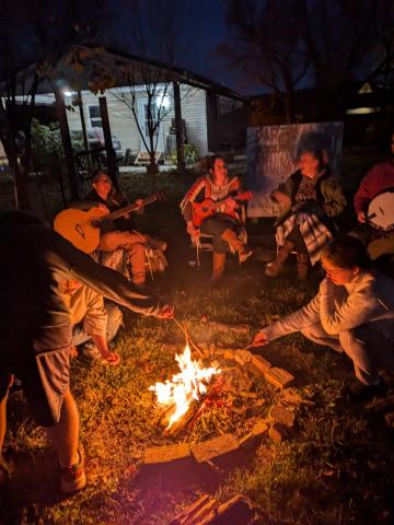 Several people laugh, sing, roast marshmallows, and play instruments around a bonfire outside Chauncey library.