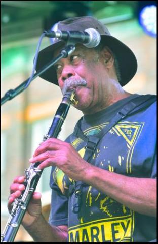 Local musician Ted Harris plays his clarinet in front of a microphone on on outdoor stage. He is wearing a Bob Marley t-shirt.