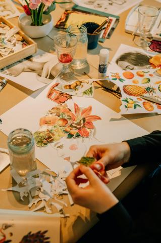 A drawing of a person's head in profile. Their hair is made of images of plants and butterflies cut out from magazines. In the foreground, a person's hands are cutting out another image of a plant. In the background, a table is covered in collage supplies.