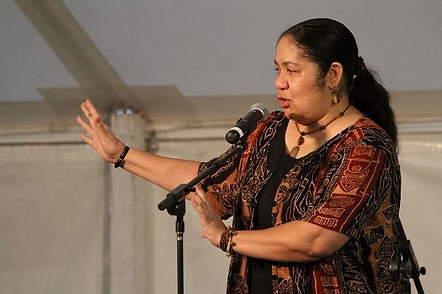 Lyn Ford speaks at a microphone. She is wearing a black and red top and has her long black hair pulled back in a ponytail.