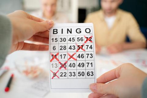 Pale, manicured hands hold a bingo card with six red X's, five of which constitute a diagonal bingo.