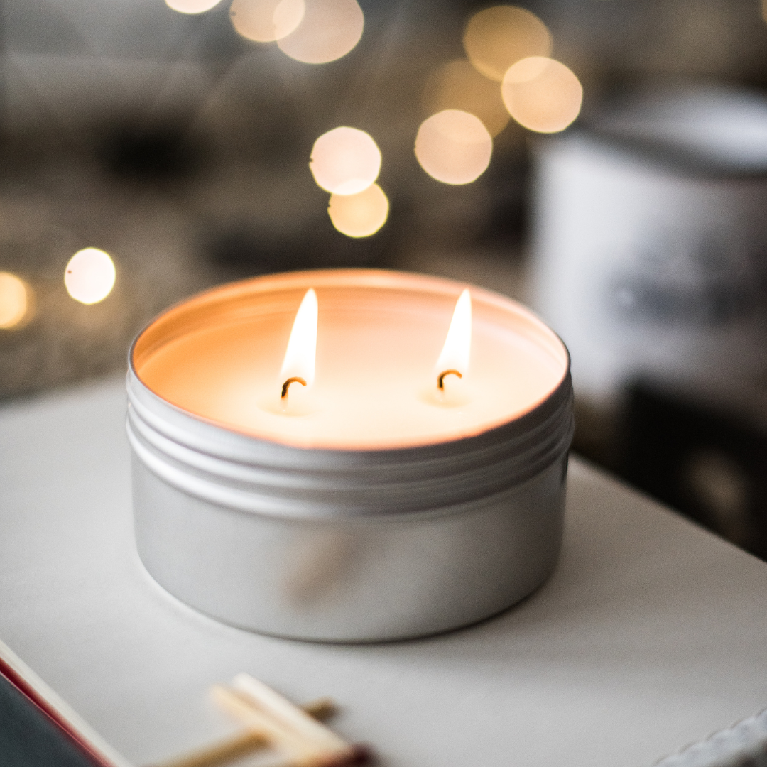 A two wick candle inside a 4 oz. silver tin sits lit atop a white surface.