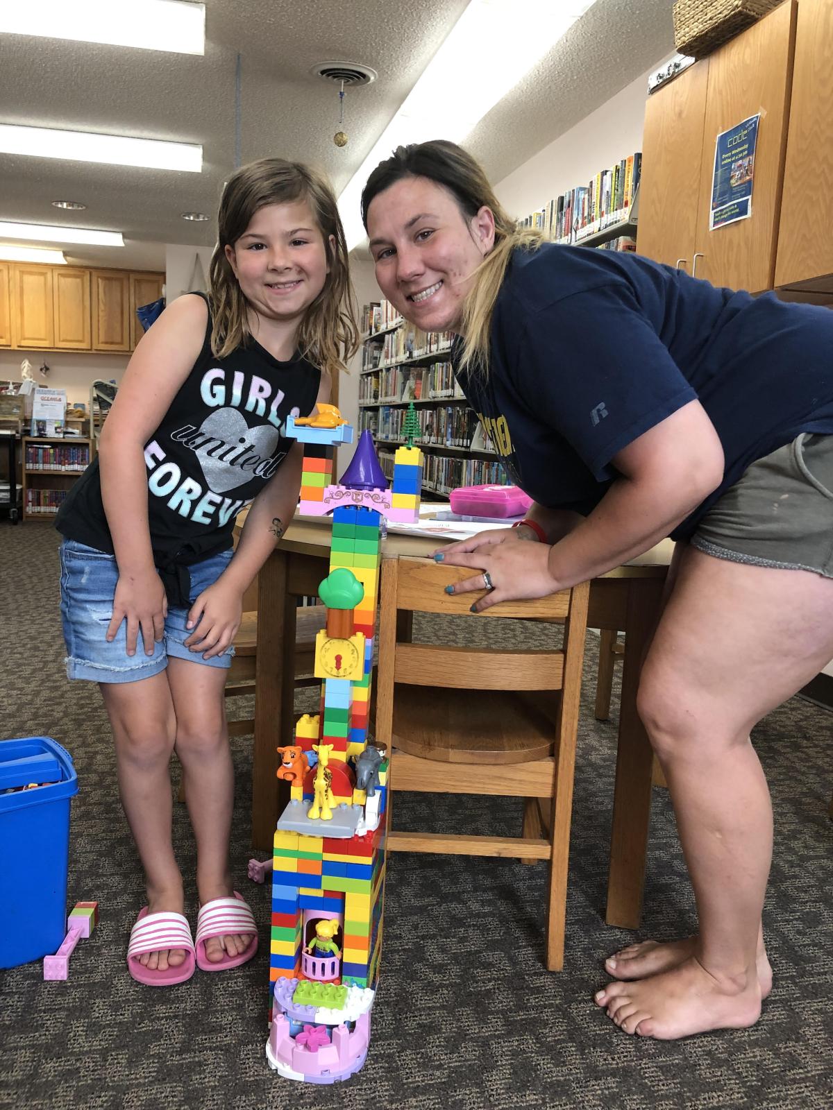 A kid and their grown-up stand on either side of an elaborate clock tower/zoo/helicopter landing pad they have constructed out of Legos.