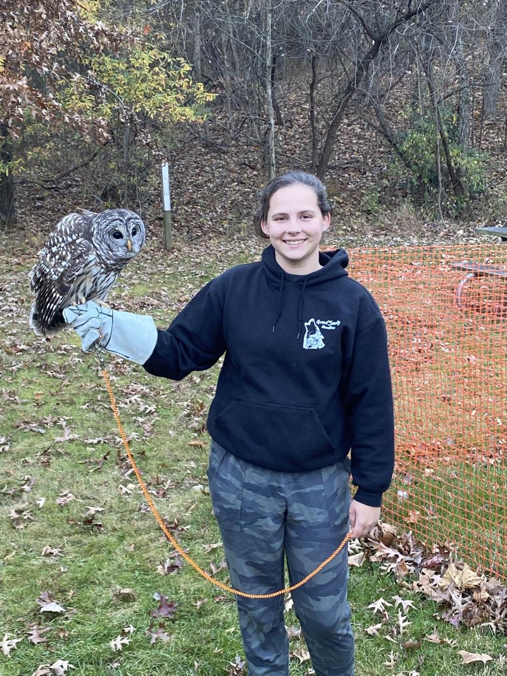 A person in a black sweatshirt and camo pants holds a barred owl on their outstreched hand.