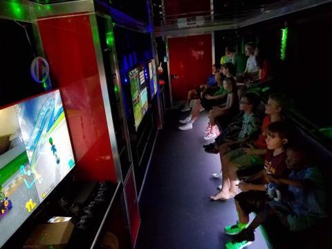 Several kids sit on benches and play video games on several big-screen TVs inside a truck