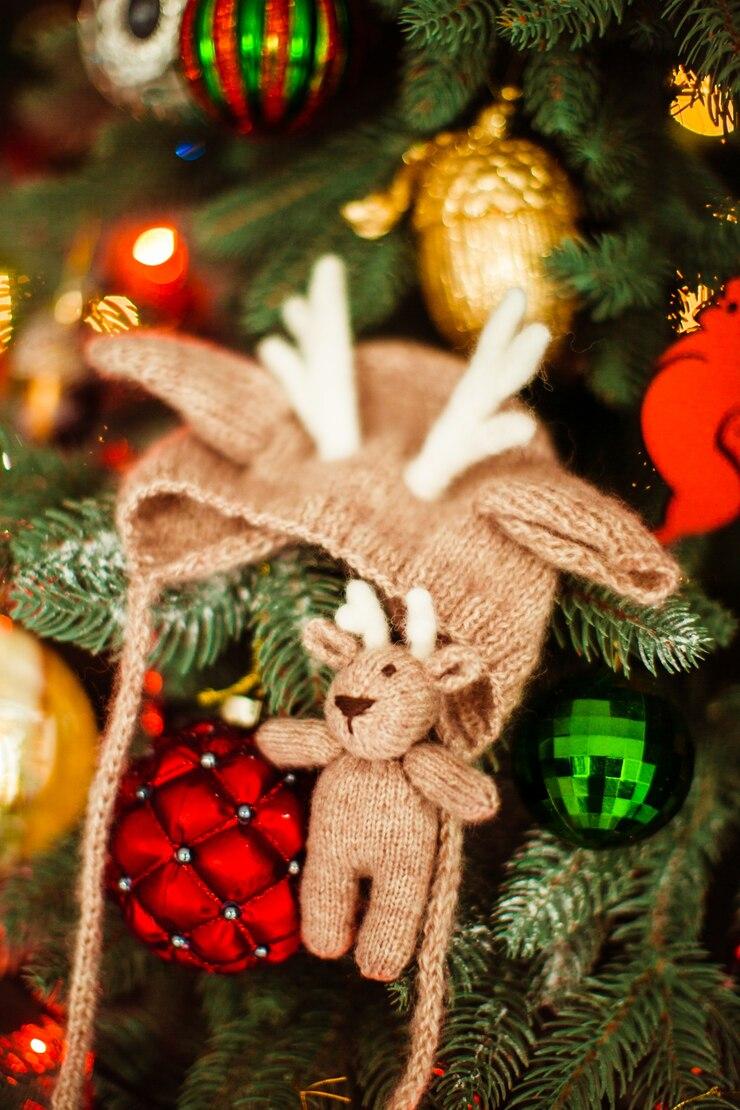 A baby hat with reindeer antlers and a knit reindeer plush hangs from a holiday tree