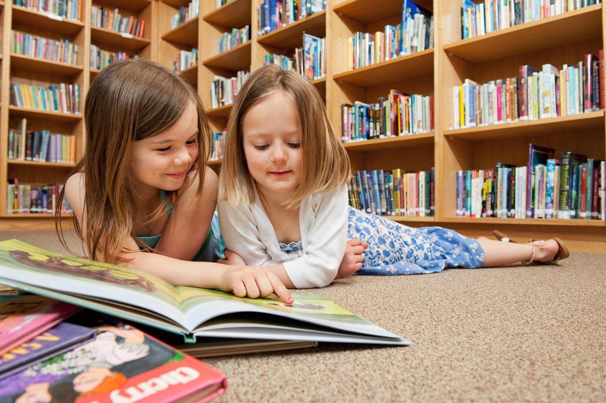 Two young children lay on their stomachs on the floor with a picture book open in front of them. 