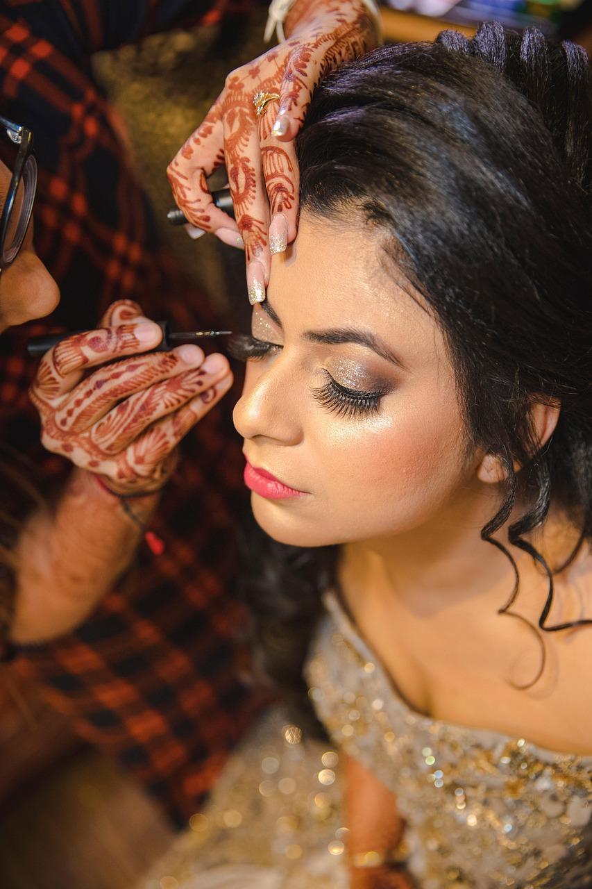 A person with henna art on their hands applies eye makeup to a person in a sequined dress.